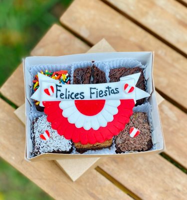 Seis postres decorados con fondant, presentados con una bandera, escarapela y corazones, empaquetados en una caja ecológica con opción de sticker personalizado.