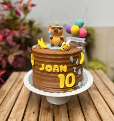 Torta decorada con buttercream, con un capibara de fondant usando lentes de sol en la parte superior, rodeado de varias bananas también en fondant. La decoración tiene un estilo divertido y tropical.