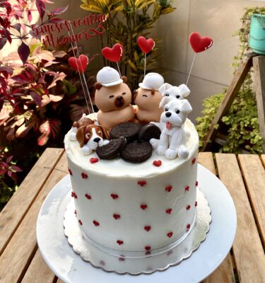 Torta decorada con dos capibaras en fondant, una dándole un beso en la mejilla a la otra, rodeadas de corazones y perritos.