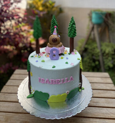 Torta decorada con una capibara en la parte superior usando tutú y gorrito de cumpleaños, rodeada de arbolitos, arbustos, hojitas y florecitas.