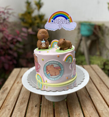 Torta decorada con dos capibaras en la parte superior, una tomando bubble tea y otra sentada, con arcoíris alrededor e imágenes de capibaras.
