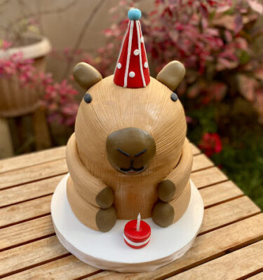 Torta en forma de capibara con gorrito de cumpleaños y un macaron con velita simulando una tortita pequeña.