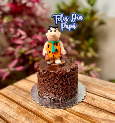 Torta decorada con trocitos de chocolate y Pedro Picapiedra en fondant sobre la parte superior, acompañada de un topper que dice “Feliz Día, Papá”.