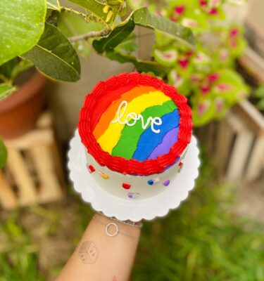 Torta temática Pride decorada con buttercream blanco, rosetones rojos y corazones con los colores de la bandera del Pride. En la parte superior lleva la palabra "Love" sobre una franja multicolor.