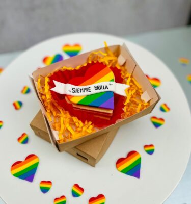 Alfajor en forma de corazón decorado con fondant en los colores de la bandera Pride y un banner con la frase “Siempre Brilla”.