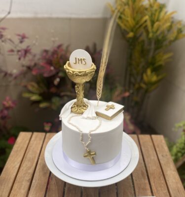 Torta de bautizo decorada con fondant, incluyendo un cáliz, rosario y biblia modelados a mano sobre la superficie.