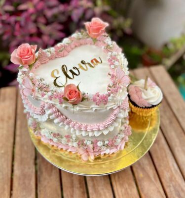 Torta en forma de corazón decorada con rosetones de buttercream en distintos tonos, flores naturales, mariposas de cartulina y un cupcake de la misma temática al costado.