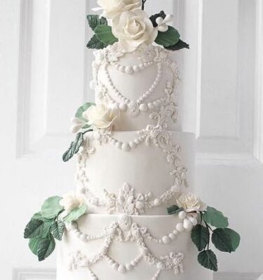 Torta de bodas de tres pisos, completamente blanca, decorada con rosetones de buttercream y coronada con rosas naturales con sus hojas verdes.