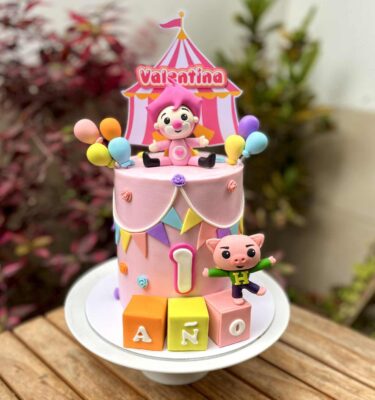 Torta de un piso en tonos rosados con temática de Plim Plim. Lleva una figura de Plim Plim rosado en la parte superior, el cerdito en la base, globos decorativos, banderines y pequeñas flores.