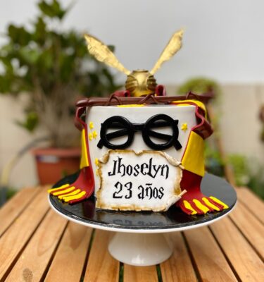 Torta temática de Harry Potter con bufanda de Gryffindor, varita mágica, lentes, Snitch dorada y pergamino personalizado en fondant.