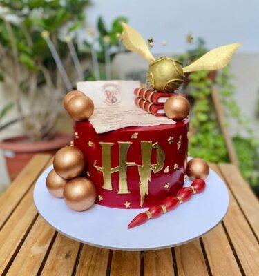 Torta de Harry Potter de un piso, forrada en fondant y decorada con elementos icónicos del mundo mágico: carta de Hogwarts, varita mágica, Snitch dorada, libros y esferas decorativas, todo elaborado en fondant.