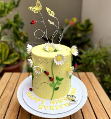 Torta bañada en buttercream, decorada con margaritas de fondant, mariquitas y mariposas de cartulina.