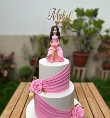 Torta de dos pisos forrada en fondant, decorada con rosas de azúcar, drapeado elegante y una muñeca en 3D de fondant de la cumpleañera, acompañada de topper dorado personalizado.