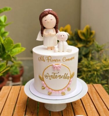 Torta de un piso para Primera Comunión, con una angelita y ovejita en 3D de azúcar, marco de fondant con flores, detalles en dorado y frase “Mi Primera Comunión”.