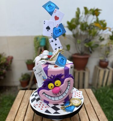 Torta de un piso de Alicia en el País de las Maravillas, forrada en fondant, con el gato sonriente en 2D al frente, el conejo en 3D en la cima y decoración de naipes en papel de arroz.