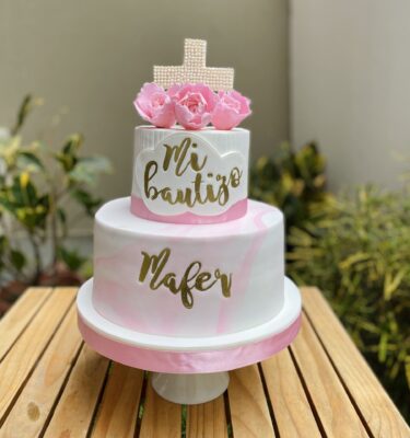 Torta de dos pisos para bautizo, decorada en fondant rosa mármol con rosas de azúcar, cruz hecha de perlitas, marco de fondant con la frase "Mi Bautizo" y nombre personalizado.
