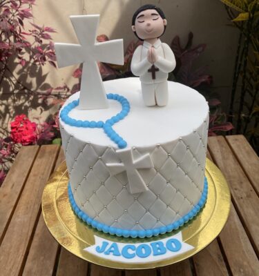 Torta de un piso con acabado capitoné en fondant, decorada con un niño modelado en 3D de azúcar, cruz y rosario en fondant, y personalización de nombre.