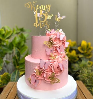 Torta de dos pisos bañada en buttercream, decorada con delicados rosetones, mariposas y un topper dorado personalizado.