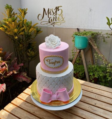 Torta de dos pisos forrada en fondant, con base en diseño capitoné, un moño decorativo alrededor, nombre personalizado en el segundo nivel y una rosa de azúcar en 3D como topper.