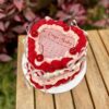 Torta Burncake en forma de corazón, decorada en tonos rojo, rosado pastel y blanco, con lazos a los lados, rosetones hechos con diferentes boquillas y cuatro cerezas brillantes en la parte superior.