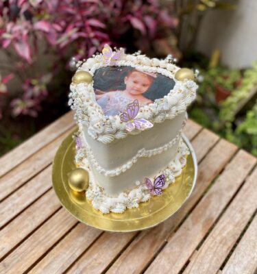 Burncake en forma de corazón, cubierto y decorado en buttercream, adornado con mariposas de cartulina, bolas de chocolate, perlas doradas comestibles y una foto del cumpleañero en el centro.