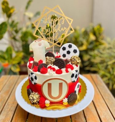Torta temática del equipo de la U bañada en buttercream blanco y rojo, decorada con pelotas de fútbol en fondant, galletas Oreo, bolas de chocolate, camiseta del equipo en papel de arroz y el símbolo del club.