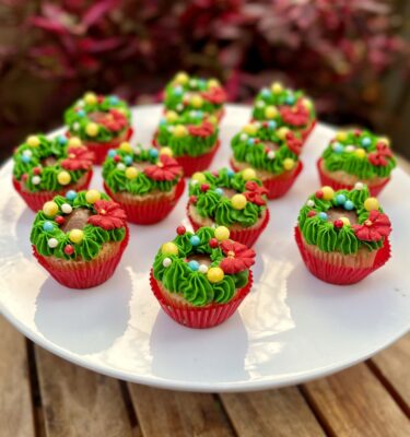 Mini cupcakes decorados con buttercream verde en forma de corona navideña, con perlitas comestibles y un lazo de fondant rojo.