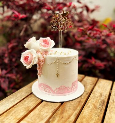 Torta de Primera Comunión con acabado en fondant blanco, encaje rosado en la parte inferior, flores de azúcar en 3D, puntitos y cruz de glaseado dorado, y topper de cartulina con la frase “Mi Primera Comunión”.