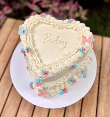 Torta en forma de corazón bañada en buttercream blanco con la palabra “baby” al centro, decorada con mariposas de papel de arroz en tonos rosado y celeste, y pequeños detalles en buttercream de los mismos colores.