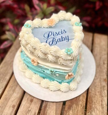 Burncake de Piscis decorado con buttercream en colores tropicales, ondas que simulan olas del mar, flores en fondant y perlas comestibles, con imagen personalizada en papel de arroz que se quema para revelar un mensaje sorpresa.