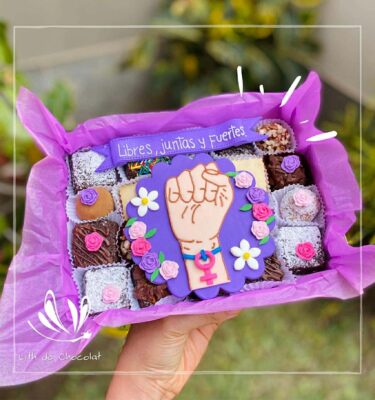 Mix box con 20 postres surtidos (brownies, alfajores y trufas) con una decoración central de puño en alto, flores en fondant y un banner que dice “Libres, juntos y fuertes”.